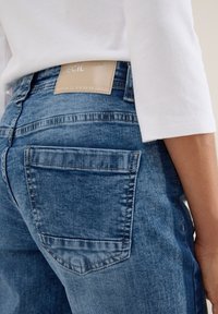 Vaqueros de mezclilla en un lavado azul descolorido con una leve textura. Incluye bolsillos traseros y una etiqueta "Cecil" de color marrón claro en la cinturilla.