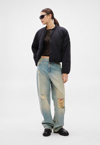 Čierna bomber bunda, skrátený strih, kombinovaná s voľnými džínsami svetlej farby s opotrebovanými detailmi. Model nosí tmavé slnečné okuliare a čierne topánky.