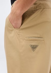 Χέρι που ξεκουράζεται στην μπροστινή τσέπη παντελονιού σε χρώμα ταμπά, με μικρή τσέπη με φερμουάρ και τρίγωνο λογότυπο PFG από κάτω.