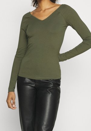 Femme portant un top vert olive à manches longues et col en V et un pantalon en cuir noir, debout avec une main sur la hanche.