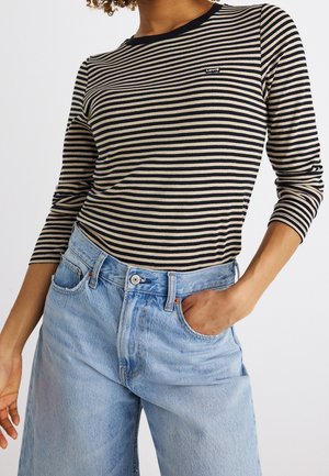 Osoba w czarno-beżowej, w paski koszuli z długim rękawem z logo Levi’s oraz jasnoniebieskich, wysokiego stanu dżinsach, ręka w kieszeni.