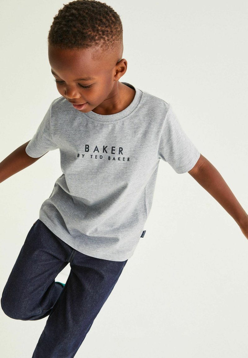 Pelēks īsām piedurknēm T-krekls ar apaļu kakla izgriezumu, priekšā ar melnu apdruku "BAKER BY TED BAKER", kombinēts ar tumši ziliem džinsiem.