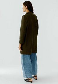 Manteau long vert olive avec un col, à coupe droite. Associé à un jean large bleu clair et des chaussures noires à enfiler.