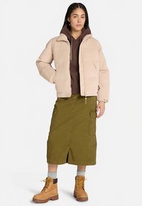 Chaqueta corta de pana beige sobre una sudadera con capucha marrón, combinada con una falda midi verde y botines marrones. Cuenta con bolsillos y detalles de cremallera.