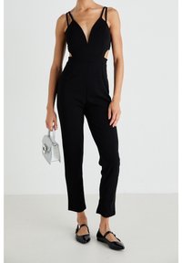 Woman wearing a black sleeveless jumpsuit with deep V-neck and cutouts, holding a small silver handbag and wearing black Mary Jane shoes.