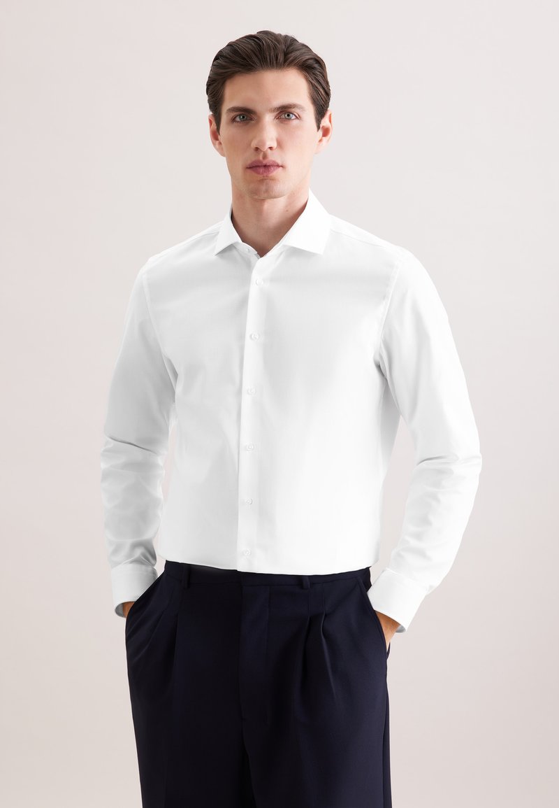 Mann steht frontal, trägt ein weißes Hemd und eine schwarze Hose, hat die Hände in den Taschen, vor einem schlichten, hellen Hintergrund.
