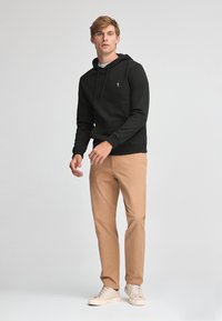 Felpa nera in cotone con coulisse, con un piccolo logo, abbinata a chinos beige chiaro e sneakers beige su uno sfondo neutro.