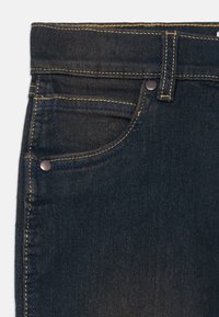 Mörkblå denimjeans med en slät yta, med gula sömmar, en framficka och en metallknapp som accent vid midjan.