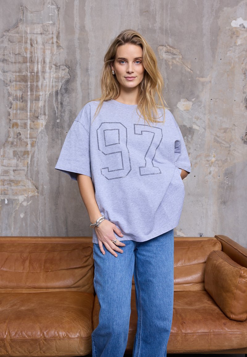 Halli üle suuruse T-särk mustade tikkudega numbriga "97" esiküljel, mille kõrvale on sobitatud helesinised laiad püksid, taustal on betoonsein.