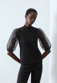 Top negru cu guler înalt, având mâneci bufante transparente. Material neted cu mansete elastice, purtat cu blugi închiși la culoare.