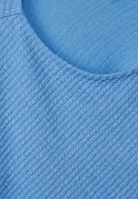 Ljusblå topp med texturerad tyg; har en slät nacklinje och vågigt mönster över materialet. Mjuk textur med en subtil lyster.