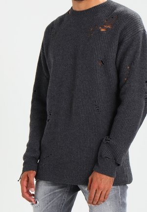 Mand iført en mørkegrå ribstrikket sweater med flere bevidste små huller og lysegrå revnede jeans.