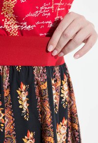 Rotes Oberteil mit goldenen Textakzenten, kombiniert mit einem gemusterten schwarzen Rock, der florale Designs in Orange, Gelb und Grün aufweist.