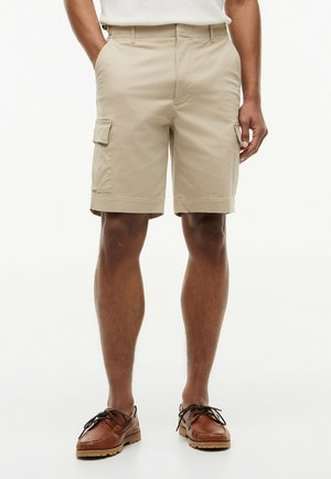 Mann trägt beige Cargo-Shorts mit Seitentaschen und braune Leder-Bootsschuhe, steht vor weißem Hintergrund.