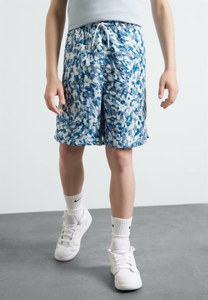 Dolna część ciała osoby noszącej szorty w kamuflażu w kolorach niebieskim, białym i szarym, białe skarpetki oraz białe trampki, stojącej na szarej podłodze.