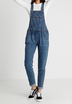 Personne portant une salopette en denim bleu avec les revers retroussés, une chemise blanche à manches longues et des baskets noires, se tenant devant un fond uni clair.