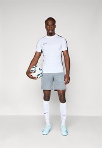 Valge lühikeste varrukatega särk mustade aktsentidega, hallid lühikesed püksid, valged sokid ja tsüaanide jalgpallijalatsid; mängija hoiab jalgpalli. Nähtav Nike bränd.