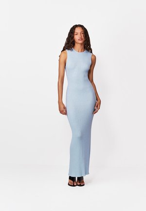 Hosszú, göndör hajú nő, aki ujjatlan, világoskék bordázott maxi ruhát és fekete nyitott orrú magassarkút visel, fehér háttér előtt áll.