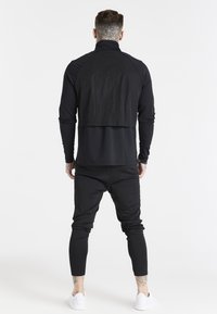 Svart atletisk topp med hög krage och raglanärmar, lagerdesign med texturerad baksida, tillsammans med svarta avsmalnande byxor och vita skor.