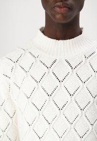 Maglione bianco lavorato a maglia con una texture a motivo diamante, dettagli a intaglio, scollatura a coste e materiale morbido e spesso.