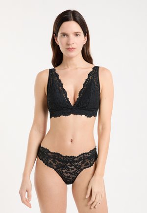 Frau trägt schwarzen Spitzen-Bralette und passende Spitzenunterwäsche, steht vor einem einfarbigen weißen Hintergrund, blickt nach vorne mit neutralem Ausdruck.