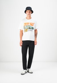 T-shirt graphique blanche avec le texte coloré "Sonic Pace", associé à un pantalon noir. Le mannequin porte un chapeau caban marine et des chaussures de sport.
