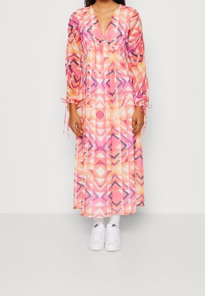 Kobieta ubrana w długą sukienkę w geometryczne wzory w odcieniach różu, pomarańczu i fioletu, do tego białe skarpetki i białe trampki.