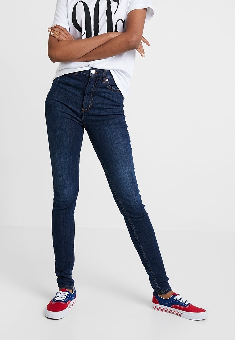 Strakke jeans in donkere denim met een hoge taille, gecombineerd met een wit T-shirt met een grote zwarte afbeelding, en rode, blauwe en witte sneakers.
