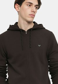 Felpa con cappuccio marrone con cerniera, caratterizzata da cordoncini neri e un piccolo logo ricamato sul petto. Texture liscia e design aderente.