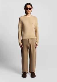 Maglione beige texture con scollatura a girocollo, abbinato a pantaloni larghi color beige. Il modello indossa occhiali da sole scuri e scarpe slip-on marroni.