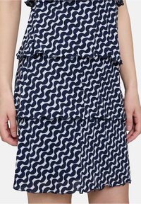 Marineblaues und weißes, geschichtetes Kleid mit einem wellenförmigen Muster, Rüschenkanten und einem leichten Stoff. Hände sind teilweise in der Taille sichtbar.