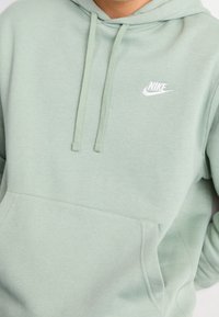 Persona vistiendo una sudadera ligera verde de Nike con bolsillo frontal y cordones, mostrando la parte superior del torso y parte del brazo.