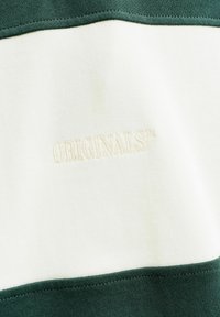 Λευκό τμήμα υφάσματος με κεντημένη λέξη "ORIGINALS" σε ανοιχτό κρεμ χρώμα, περιβαλλόμενο από σκοτεινές πράσινες υφασμάτινες λωρίδες στην κορυφή και στο κάτω μέρος.