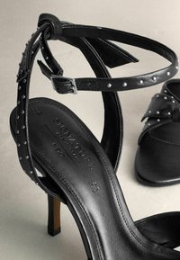 Schwarze High-Heel-Sandalen mit genagelten Riemen, aus glattem Leder und einem schlanken Knöchelverschluss. Sie verfügen über einen rechteckigen Blockabsatz.