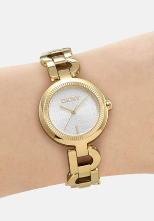 Goudkleurig DKNY polshorloge met witte wijzerplaat met patroon en schakelsluiting om de pols van een persoon, klok wijst 3:10:34 aan.