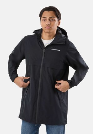 Młody mężczyzna w czarnej kurtce z kapturem i zamkiem błyskawicznym, poprawiający kieszenie, stoi na tle jednolitego białego tła.