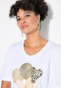 Camiseta de algodón blanca con un aplique de corazón multicolor con patrones, incluyendo rayas y print de leopardo, acentuado con tachuelas doradas.
