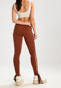 Mujer vistiendo leggings marrones con detalles de cordones laterales, sujetador deportivo beige y zapatillas de plataforma blancas, de pie con la espalda hacia la cámara.