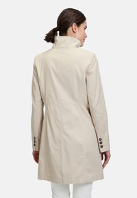 Beige Mantel mit hohem Kragen, Seitentaschen und sechs Knopf-Details an den Ärmeln. Glatte Textur, taillierte Passform, hüftlanger Schnitt.