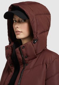 Chaqueta acolchada burdeos con capucha, que dispone de cremallera y botones a presión, y una gorra negra, que muestra una textura suave y acolchada.