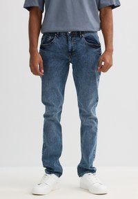 Vaqueros de denim azul con un ajuste recto, que cuentan con cinco bolsillos, un desgaste sutil y detalles de costura. Combinados con zapatillas blancas y una camiseta gris.