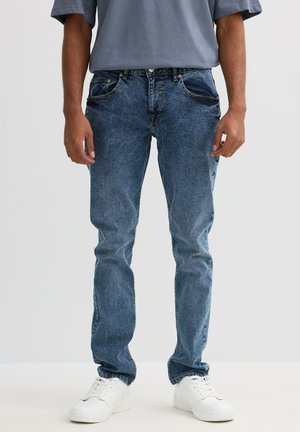 Vaqueros de denim azul con un ajuste recto, que cuentan con cinco bolsillos, un desgaste sutil y detalles de costura. Combinados con zapatillas blancas y una camiseta gris.