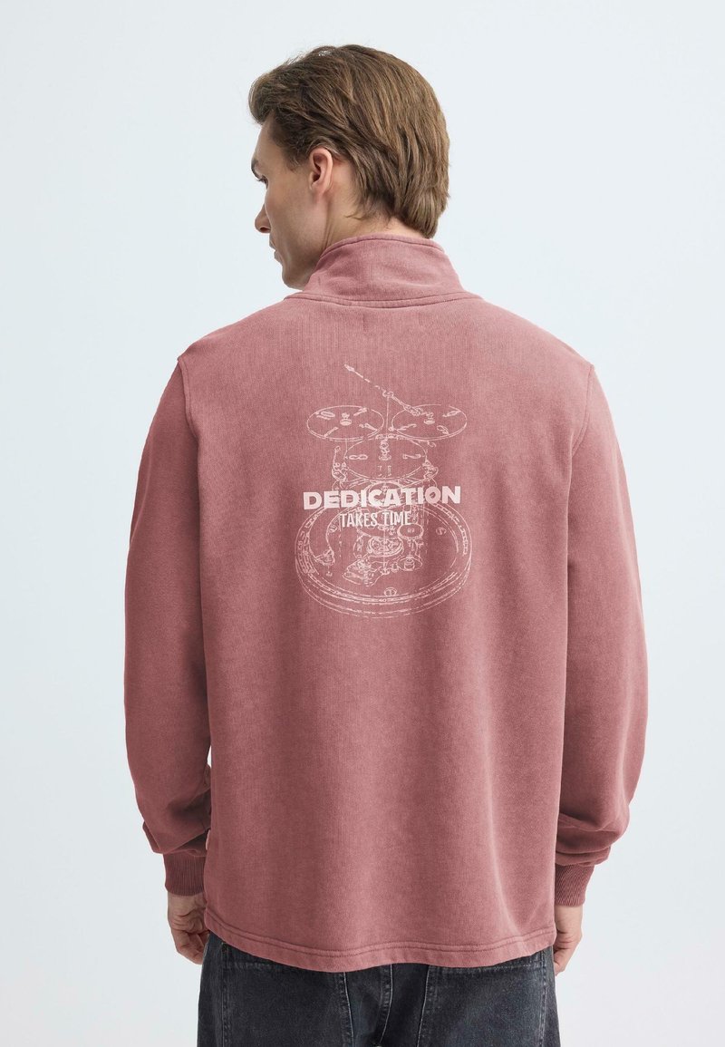 Roze pullover met hoge kraag, met een witte grafische print op de achterkant met de tekst "DEDICATION TAKES TIME." Zachte textuur.