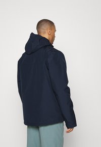 Chaqueta impermeable azul marino con capucha, que presenta una textura suave y un corte recto. Sin logotipos visibles; la vista trasera muestra un diseño simple.