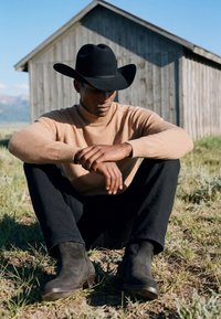 Homme portant un chapeau de cow-boy noir, un pull beige, un pantalon noir et des bottes, assis en tailleur sur l’herbe près d'une grange en bois sous un ciel dégagé.