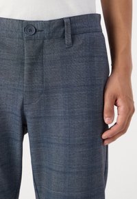 Dunkelblaue Karohosen mit einem Knopfverschluss, die eine glatte Textur und eine taillierte Passform aufweisen. Die Hand ist seitlich zur Veranschaulichung positioniert.