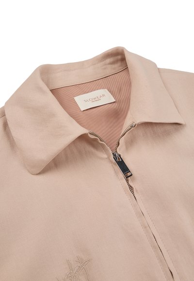 Beige collared jacket with front zipper partially open, featuring a "SLOWEAR" label inside the collar and subtle embroidered design near bottom left.