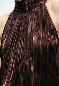 Κοντινή λήψη μεταλλικού μπρονζέ πλισέ αμάνικου τοπ με ψηλό λαιμό σε φόντο ανοιχτού δέρματος.