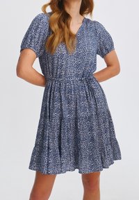 Kobieta w niebieskiej sukience z krótkim rękawem, ozdobionej białym kwiatowym wzorem, z dekoltem w kształcie litery V, zebranym w talii i rozkloszowanym dołem, z rękami opartymi na biodrach.