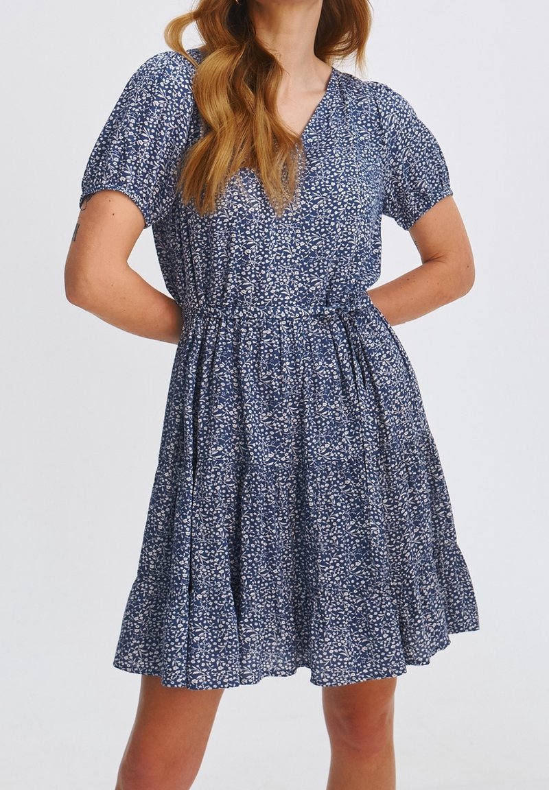 Kobieta w niebieskiej sukience z krótkim rękawem, ozdobionej białym kwiatowym wzorem, z dekoltem w kształcie litery V, zebranym w talii i rozkloszowanym dołem, z rękami opartymi na biodrach.